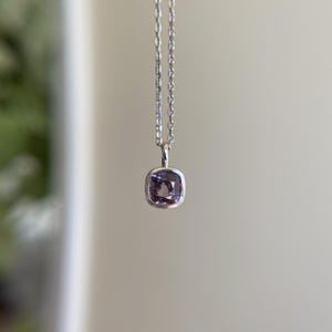 May include: A silver necklace with a square, light purple gemstone pendant. The pendant is set in a silver bezel and hangs from a delicate silver chain. The background is a soft, neutral color.