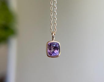 May include: A gold necklace featuring a square, purple gemstone pendant. The pendant is set in a gold bezel and suspended from a delicate chain. The gemstone has a faceted surface, reflecting light.