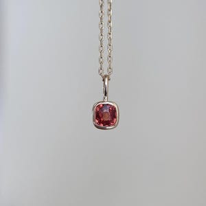 May include: A delicate pendant necklace featuring a square, reddish-pink gemstone set in a gold-colored metal. The gemstone is suspended from a fine chain, creating a minimalist and elegant design. The necklace is a piece of jewelry.