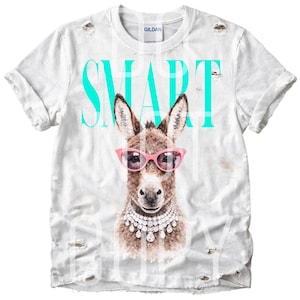May include: White distressed t-shirt featuring a watercolor illustration of a donkey wearing pink sunglasses and a diamond necklace. The word "SMART" is printed in large, turquoise letters across the top.