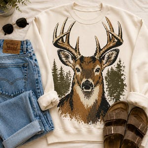 Pode incluir: Uma camisola de gola redonda cor de creme com um grande gráfico de uma cabeça de veado castanha com hastes, centrada contra uma silhueta de floresta verde escura. A camisola é combinada com calças de ganga azuis, óculos de sol pretos e sandálias castanhas.