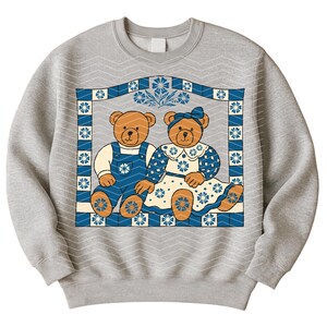 May include: A gray sweatshirt featuring a graphic of two teddy bears within a blue and white floral frame. One bear wears blue overalls, the other a blue dress. The background has a repeating chevron pattern.