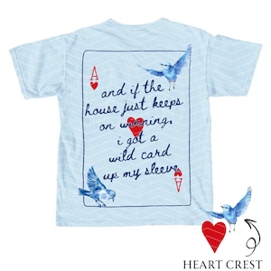 May include: Light blue t-shirt with a white chevron pattern. The back of the shirt features a playing card design with text and illustrations of blue birds. The text reads "and if the house just keeps on winning, I got a wild card up my sleeve."