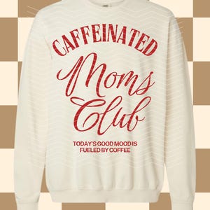 Puede incluir: Sudadera color crema con la frase "CAFFEINATED Moms Club" en rojo. Debajo, se lee "TODAY'S GOOD MOOD IS FUELED BY COFFEE". La sudadera tiene cuello redondo y mangas largas.