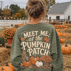 Puede incluir: Una sudadera de manga larga de color verde oliva con un gráfico en la espalda que incluye el texto "MEET ME at the PUMPKIN PATCH" en letras color crema, rodeado de ilustraciones de calabazas naranjas, una calabaza blanca y follaje otoñal.