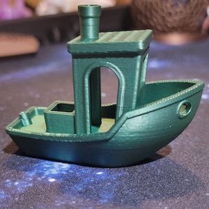 May include: A small, green, 3D printed model of a boat with a chimney. The boat has a rounded hull and a small cabin with a window.