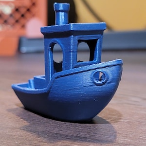Könnte beinhalten: Ein kleines, blaues, 3D-gedrucktes Spielzeugboot mit einem Schornstein. Das Boot hat einen abgerundeten Rumpf und eine kleine Kabine mit einem Fenster.