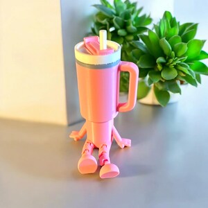 May include: A pink plastic toy shaped like a cup with a straw and a handle. The cup has a white lid and a gray band around the middle. The toy has arms and legs and is sitting on a gray surface.