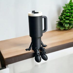 May include: A black 3D printed mug with a white rim and a handle sits on a wooden surface. The mug has two black legs and arms, giving it a whimsical, cartoon-like appearance.
