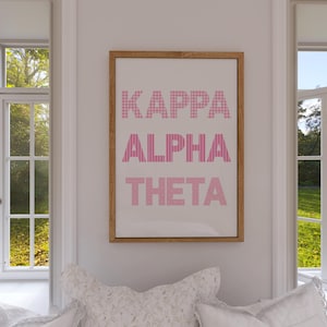 May include: A framed print with the Greek letters Kappa, Alpha, and Theta in pink. The letters are displayed vertically on a white background. The frame is a light brown color. The print is hanging on a white wall between two windows.