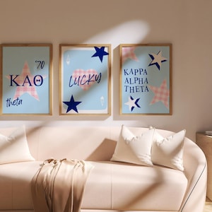 May include: Three framed prints with light blue backgrounds, wooden frames, and various designs. The prints feature text and star shapes in pink and blue. A sofa with white cushions and a throw is in the foreground.