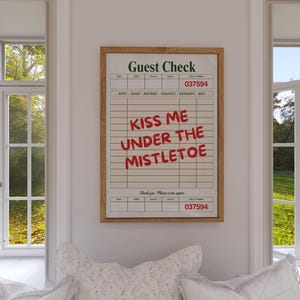 May include: A framed print with the words "Kiss Me Under The Mistletoe" in red on a vintage-style guest check. The print is in a light brown wooden frame and hangs on a white wall between two windows.