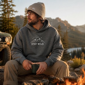Peut inclure: Un sweat à capuche gris avec le texte "STAY WILD" et un motif de montagne. La personne porte un bonnet beige et tient une tasse verte. L'arrière-plan comprend un véhicule, des arbres et des montagnes.