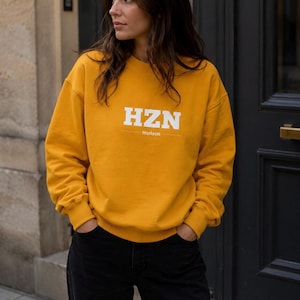 Peut inclure: Sweat-shirt jaune moutarde avec le texte blanc "HZN" et "Horizon" en dessous. Le sweat-shirt est de style col rond et est associé à un pantalon noir. Le modèle se tient devant une porte sombre.