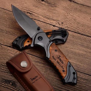 Könnte beinhalten: Ein Klappmesser mit einer dunkelgrauen Klinge und einem Holzgriff mit der Gravur Jason Happy Father's Day, begleitet von einer passenden braunen Lederscheide, die ebenfalls mit Jason Happy Father's Day graviert ist.