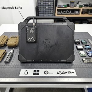 May include: A black Dell laptop with a magnetic LoRa device attached. The device has an antenna and the text "Magnetic LoRa". Other items on the table include small electronic components, a multi-tool, and pouches. The table also has a ruler.