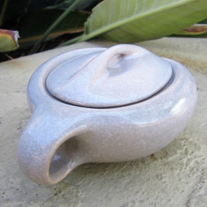 May include: A light gray ceramic teapot with a matching lid. The teapot has a rounded body, a curved handle, and a spout. The lid has a loop handle. The teapot is sitting on a textured surface.