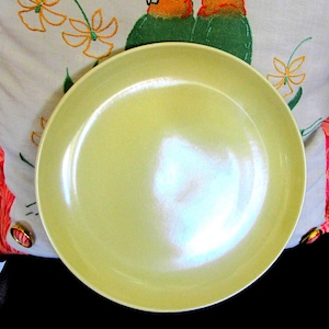 May include: A pale yellow ceramic plate with a glossy finish. The plate is round with a slightly raised edge. The background includes an embroidered pillow with orange and green bird designs and floral patterns.