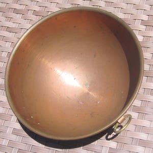 K&ouml;nnte beinhalten: Eine Kupfersch&uuml;ssel mit Messingrand und kleinem Messingringgriff. Die Sch&uuml;ssel hat eine abgerundete Form und eine polierte Oberfl&auml;che, die das Licht reflektiert. Das Kupfer hat einen warmen, r&ouml;tlich-braunen Farbton, was auf einen antiken oder Vintage-Artikel hindeutet.