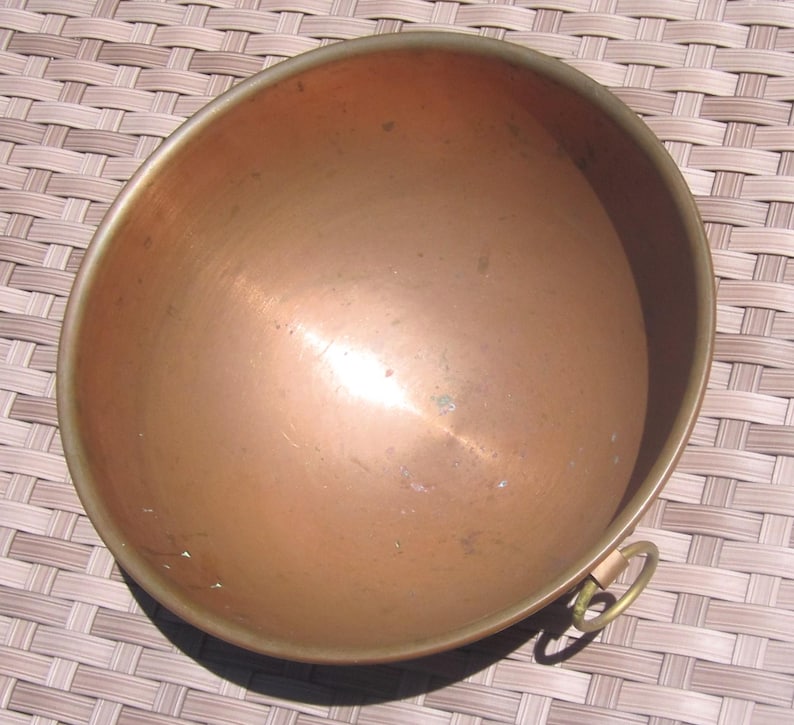 K&ouml;nnte beinhalten: Eine Kupfersch&uuml;ssel mit Messingrand und kleinem Messingringgriff. Die Sch&uuml;ssel hat eine abgerundete Form und eine polierte Oberfl&auml;che, die das Licht reflektiert. Das Kupfer hat einen warmen, r&ouml;tlich-braunen Farbton, was auf einen antiken oder Vintage-Artikel hindeutet.