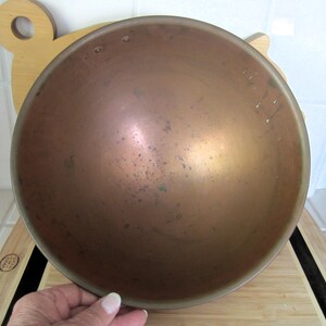 K&ouml;nnte beinhalten: Eine kupferfarbene Sch&uuml;ssel mit runder Form. Die Sch&uuml;ssel hat ein leicht angelaufenes Aussehen mit dunkleren Flecken und Variationen auf der Metalloberfl&auml;che. Die Sch&uuml;ssel ist wahrscheinlich aus Kupfer oder einer Kupferlegierung. Die Sch&uuml;ssel steht auf einem Holzschneidebrett.