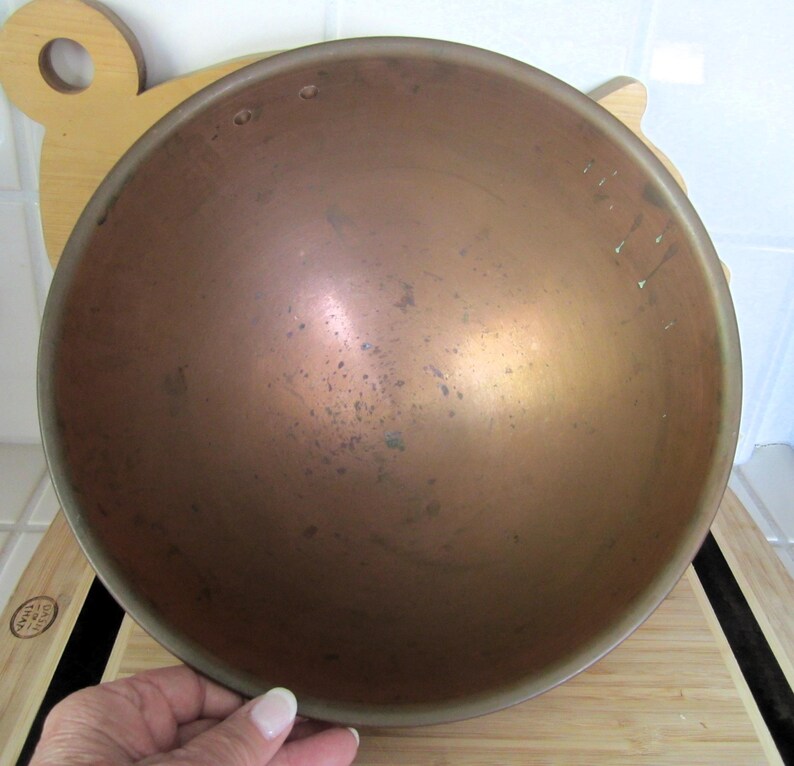 K&ouml;nnte beinhalten: Eine kupferfarbene Sch&uuml;ssel mit runder Form. Die Sch&uuml;ssel hat ein leicht angelaufenes Aussehen mit dunkleren Flecken und Variationen auf der Metalloberfl&auml;che. Die Sch&uuml;ssel ist wahrscheinlich aus Kupfer oder einer Kupferlegierung. Die Sch&uuml;ssel steht auf einem Holzschneidebrett.