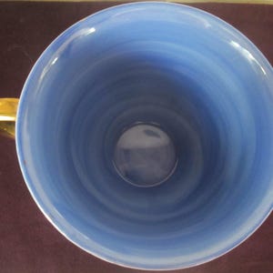 K&ouml;nnte beinhalten: Draufsicht auf eine Keramik-Teetasse mit hellblauem Innenraum und einem goldfarbenen Henkel. Das Innere der Tasse weist ein wirbelndes Muster aus hellen und dunklen Blaut&ouml;nen auf, das ein strukturiertes Aussehen erzeugt. Der Henkel ist goldfarben gl&auml;nzend.