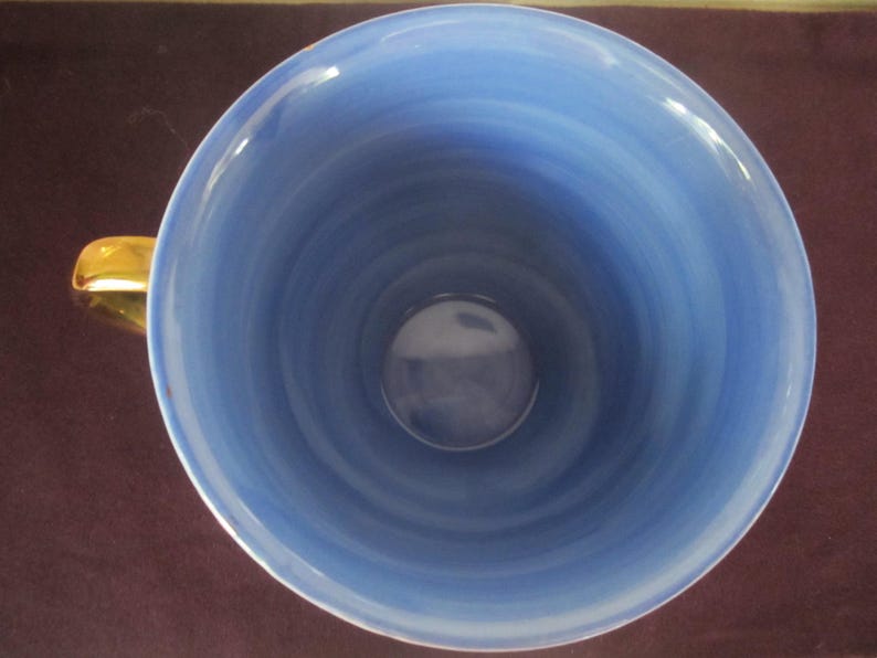 K&ouml;nnte beinhalten: Draufsicht auf eine Keramik-Teetasse mit hellblauem Innenraum und einem goldfarbenen Henkel. Das Innere der Tasse weist ein wirbelndes Muster aus hellen und dunklen Blaut&ouml;nen auf, das ein strukturiertes Aussehen erzeugt. Der Henkel ist goldfarben gl&auml;nzend.
