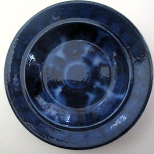 May include: A deep blue ceramic plate with a glossy finish. The plate has a circular design with a raised outer rim and a central recessed area. The color is a rich, dark blue with lighter blue accents, creating a marbled effect.