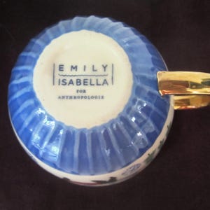 K&ouml;nnte beinhalten: Eine Keramik-Teetasse mit blau-wei&szlig;em Rippendesign. Der Griff ist goldfarben. Auf dem Boden der Tasse steht der Text "EMILY ISABELLA FOR ANTHROPOLOGIE".