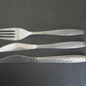 May include: A set of three stainless steel utensils: a fork, a knife, and a butter knife. The fork has four tines. The knife and butter knife have curved blades. All three utensils have textured handles.