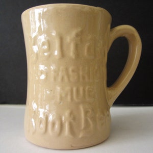 May include: Tan ceramic mug with a curved handle. The mug has embossed text that reads "Built for Fashion Mug, Look Real". The mug is cylindrical and has a slightly flared top.