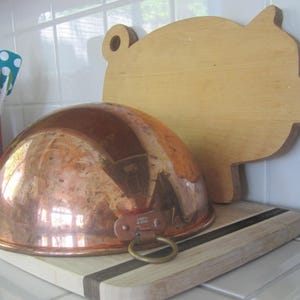 K&ouml;nnte beinhalten: Eine Kupfersch&uuml;ssel mit Messingringgriff liegt auf einem Holzschneidebrett mit schwarzem Streifen. Ein schweinef&ouml;rmiges Holzschneidebrett befindet sich im Hintergrund. K&uuml;chenutensilien sind in einem roten Beh&auml;lter links.