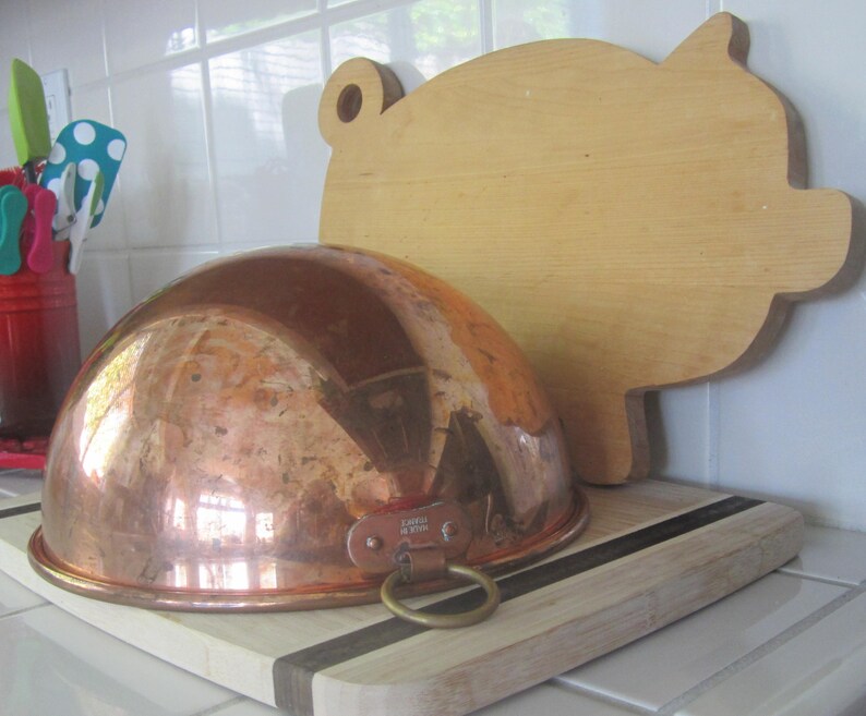 K&ouml;nnte beinhalten: Eine Kupfersch&uuml;ssel mit Messingringgriff liegt auf einem Holzschneidebrett mit schwarzem Streifen. Ein schweinef&ouml;rmiges Holzschneidebrett befindet sich im Hintergrund. K&uuml;chenutensilien sind in einem roten Beh&auml;lter links.