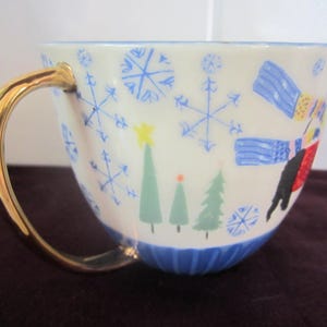 K&ouml;nnte beinhalten: Eine wei&szlig;e Keramik-Teetasse mit goldenem Henkel, verziert mit blauen Schneeflocken, gr&uuml;nen Weihnachtsb&auml;umen und einem Skifahrer. Der Boden der Tasse ist blau.