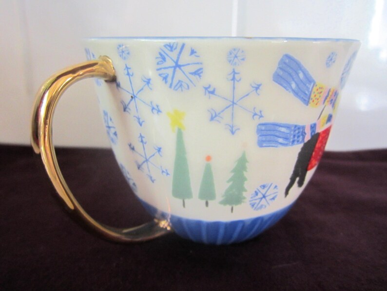 K&ouml;nnte beinhalten: Eine wei&szlig;e Keramik-Teetasse mit goldenem Henkel, verziert mit blauen Schneeflocken, gr&uuml;nen Weihnachtsb&auml;umen und einem Skifahrer. Der Boden der Tasse ist blau.