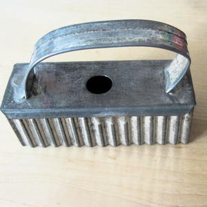 May include: A vintage rectangular cookie cutter with a corrugated edge and a handle. The metal cutter has a dark, aged appearance, with a small hole in the center. The handle is arched and attached to the top of the cutter.
