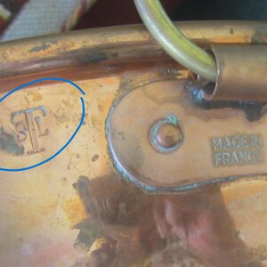 K&ouml;nnte beinhalten: Nahaufnahme eines Kupfertopfes mit Messinggriff und -schlaufe. Der Topf hat ein angelaufenes Aussehen mit dem Text "MADE IN FRANCE" auf der Seite. Es gibt auch eine "STF"-Markierung auf der Seite.