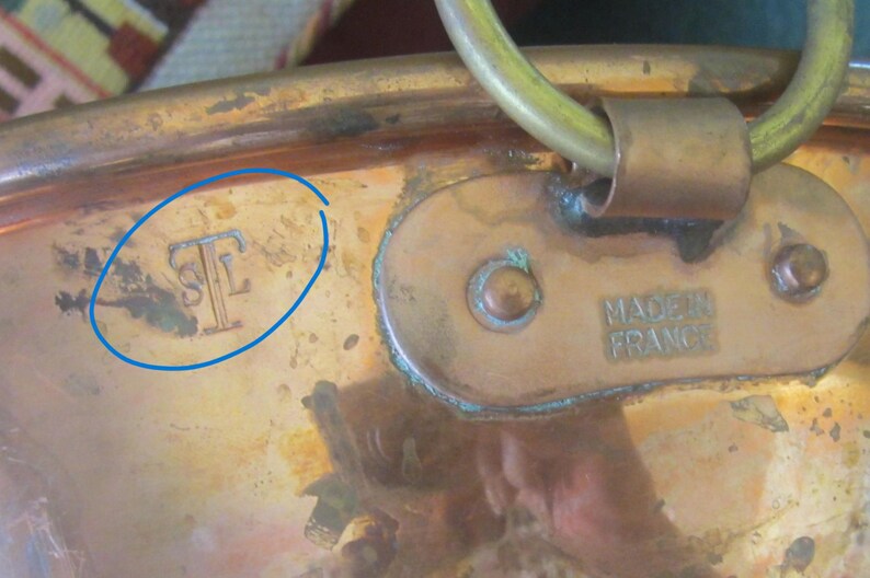K&ouml;nnte beinhalten: Nahaufnahme eines Kupfertopfes mit Messinggriff und -schlaufe. Der Topf hat ein angelaufenes Aussehen mit dem Text "MADE IN FRANCE" auf der Seite. Es gibt auch eine "STF"-Markierung auf der Seite.