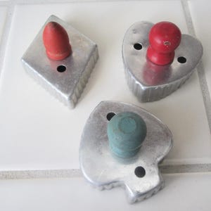 May include: Three vintage metal cookie cutters in diamond, heart, and spade shapes. Each cutter has a wooden knob handle in red or teal. The cutters are on a white tiled surface.