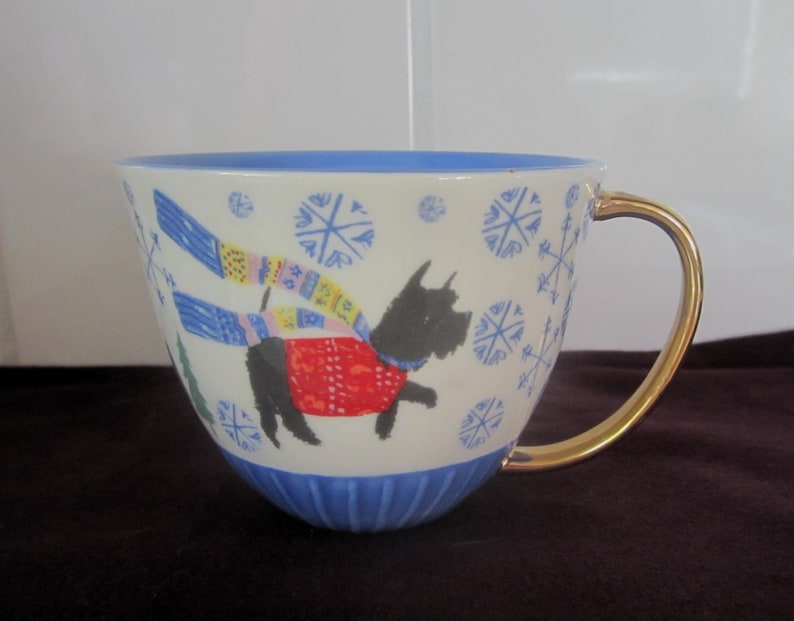 K&ouml;nnte beinhalten: Eine wei&szlig;e Keramiktasse mit blauem Rand und Boden, die einen schwarzen Hund mit rotem Pullover und buntem Schal zeigt. Die Tasse hat einen goldenen Henkel und Schneeflockenmuster.