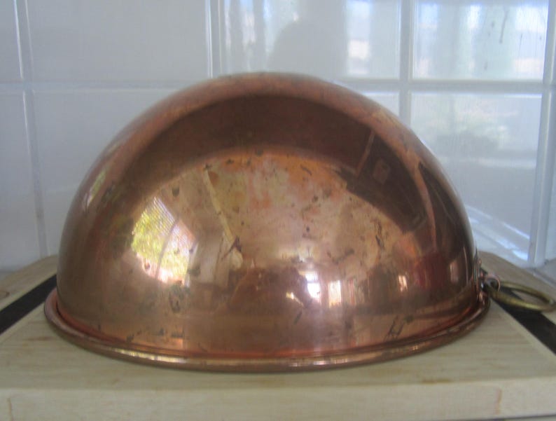 K&ouml;nnte beinhalten: Eine gro&szlig;e Kupfersch&uuml;ssel mit einer abgerundeten, kuppelf&ouml;rmigen Form. Die Sch&uuml;ssel hat eine gl&auml;nzende, reflektierende Oberfl&auml;che mit einigen Verf&auml;rbungen. Ein kleiner Messingring ist an der Seite befestigt. Die Sch&uuml;ssel steht auf einer Holzoberfl&auml;che.