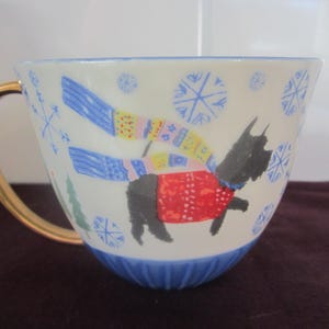 K&ouml;nnte beinhalten: Eine wei&szlig;e Keramik-Tasse mit goldenem Henkel und blauem Boden. Die Tasse zeigt eine skurrile Illustration eines schwarzen Hundes mit rotem Pullover und blau-gelbem Schal, mit Schneeflocken und einem Weihnachtsbaum-Design.