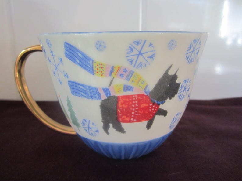 K&ouml;nnte beinhalten: Eine wei&szlig;e Keramik-Tasse mit goldenem Henkel und blauem Boden. Die Tasse zeigt eine skurrile Illustration eines schwarzen Hundes mit rotem Pullover und blau-gelbem Schal, mit Schneeflocken und einem Weihnachtsbaum-Design.