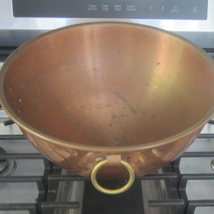 K&ouml;nnte beinhalten: Eine Kupfersch&uuml;ssel mit runder Form und einem goldenen Ringgriff. Die Sch&uuml;ssel steht auf einem Gasherd. Das Kupfer hat eine warme, r&ouml;tlich-braune Farbe.