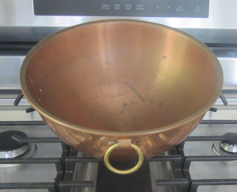 K&ouml;nnte beinhalten: Eine Kupfersch&uuml;ssel mit runder Form und einem goldenen Ringgriff. Die Sch&uuml;ssel steht auf einem Gasherd. Das Kupfer hat eine warme, r&ouml;tlich-braune Farbe.