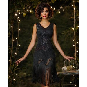 May include: A woman wearing a black sequined flapper dress with fringe. The dress has a V-neckline and is sleeveless. The woman is standing in front of a backdrop of greenery and lights.