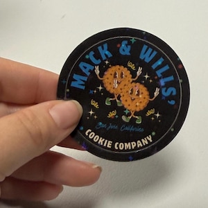 Könnte beinhalten: Ein runder, schwarzer Aufkleber mit dem Text "MACK & WILLS" in Blau, der zwei animierte Kekse mit Armen und Beinen zeigt. Der Aufkleber enthält auch den Text "San Jose, California" und "COOKIE COMPANY".