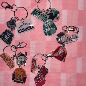 May include: A collection of hockey-themed keychains featuring team logos and jersey designs. Keychains include the Vancouver Canucks, San Jose Sharks, Anaheim Ducks, and New Jersey Devils. Each keychain has a silver keyring and clasp.