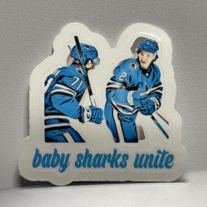 Könnte beinhalten: Ein Aufkleber mit zwei Eishockeyspielern in blauen Uniformen. Der Text "baby sharks unite" ist hellblau. Die Spieler sind in Aktion und halten Hockeyschläger.